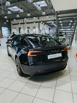 A sleek, black electric car is positioned on a tiled showroom floor. The modern interior of the showroom features large windows, numerous overhead lights, and industrial-style metal ceiling structures. Other vehicles and showroom elements can be seen in the background.