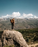 A person stands on a rocky ledge, gazing out at a vast landscape of rolling hills and distant snow-capped mountains under a bright blue sky adorned with fluffy white clouds. The individual is wearing a brown jacket and a large orange backpack, conveying a sense of exploration and adventure.
