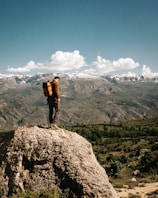 A person stands on a rocky ledge, gazing out at a vast landscape of rolling hills and distant snow-capped mountains under a bright blue sky adorned with fluffy white clouds. The individual is wearing a brown jacket and a large orange backpack, conveying a sense of exploration and adventure.