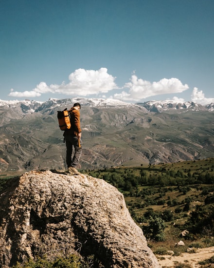 A person stands on a rocky ledge, gazing out at a vast landscape of rolling hills and distant snow-capped mountains under a bright blue sky adorned with fluffy white clouds. The individual is wearing a brown jacket and a large orange backpack, conveying a sense of exploration and adventure.
