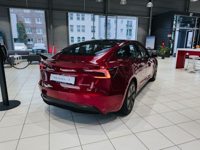 A red electric car is parked inside a modern showroom with large windows. The car's license plate reads 'Model 3' and it is a dual motor variant. The showroom has a sleek interior design with white tiled floors and seating areas visible in the background.