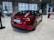 A red electric car is parked inside a modern showroom with large windows. The car's license plate reads 'Model 3' and it is a dual motor variant. The showroom has a sleek interior design with white tiled floors and seating areas visible in the background.