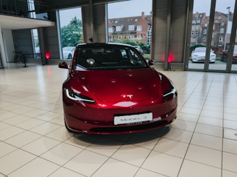 A red Tesla Model 3 is displayed in a spacious and modern showroom with large windows, white tiled flooring, and a 'Happy Halloween' sign visible outside. Several buildings and other cars are seen through the windows.