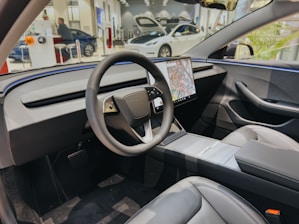 A modern car interior features a sleek steering wheel with a large central touchscreen displaying a map. The dashboard is minimalistic and futuristic, with smooth lines and integrated controls. The seats appear comfortable, made from a high-quality material, and the overall design is clean and uncluttered.