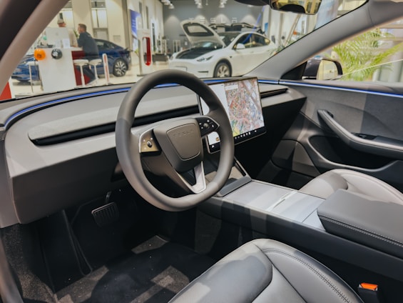A modern car interior features a sleek steering wheel with a large central touchscreen displaying a map. The dashboard is minimalistic and futuristic, with smooth lines and integrated controls. The seats appear comfortable, made from a high-quality material, and the overall design is clean and uncluttered.