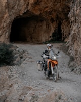 A motorcyclist wearing protective gear rides an orange adventure motorcycle on a rocky path leading into a large cave. The environment is rugged, with rocky walls and sparse vegetation.