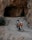 A motorcyclist wearing protective gear rides an orange adventure motorcycle on a rocky path leading into a large cave. The environment is rugged, with rocky walls and sparse vegetation.