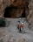 A motorcyclist wearing protective gear rides an orange adventure motorcycle on a rocky path leading into a large cave. The environment is rugged, with rocky walls and sparse vegetation.