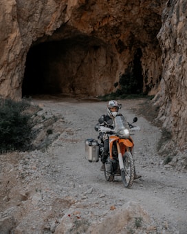 A motorcyclist wearing protective gear rides an orange adventure motorcycle on a rocky path leading into a large cave. The environment is rugged, with rocky walls and sparse vegetation.