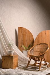 A cozy interior setup featuring a rattan chair placed on a woven rug. There are two potted plants, one on a wooden cube and another on the floor. A wooden folding screen and soft white drapery complete the background, creating a serene and natural atmosphere.
