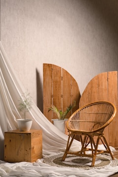 A cozy interior setup featuring a rattan chair placed on a woven rug. There are two potted plants, one on a wooden cube and another on the floor. A wooden folding screen and soft white drapery complete the background, creating a serene and natural atmosphere.
