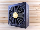 A black and gold power supply unit featuring a prominent cooling fan with a circular grille and a gold emblem in the center. The surrounding wood-textured surface accentuates the industrial design of the unit. The brand name is prominently displayed on the side and on the fan grille.