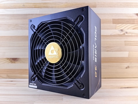 A black and gold power supply unit featuring a prominent cooling fan with a circular grille and a gold emblem in the center. The surrounding wood-textured surface accentuates the industrial design of the unit. The brand name is prominently displayed on the side and on the fan grille.