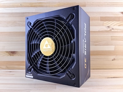 A black and gold power supply unit featuring a prominent cooling fan with a circular grille and a gold emblem in the center. The surrounding wood-textured surface accentuates the industrial design of the unit. The brand name is prominently displayed on the side and on the fan grille.
