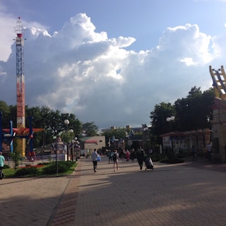 A theme park scene featuring an open walkway lined with trees and lampposts. People walk along the path, some pulling luggage. A colorful, tall tower ride is visible to the left, and another amusement structure is partially visible to the right. Large, fluffy clouds fill the sky, casting some shadows on the ground.