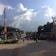 A theme park scene featuring an open walkway lined with trees and lampposts. People walk along the path, some pulling luggage. A colorful, tall tower ride is visible to the left, and another amusement structure is partially visible to the right. Large, fluffy clouds fill the sky, casting some shadows on the ground.