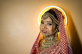 A bride adorned in traditional attire with intricate gold jewelry, including a nose ring, earrings, and a heavily embroidered red dupatta. Her makeup is elegantly done, featuring a bindi on her forehead. A halo-like circular light creates a soft glow behind her head.