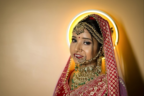 A bride adorned in traditional attire with intricate gold jewelry, including a nose ring, earrings, and a heavily embroidered red dupatta. Her makeup is elegantly done, featuring a bindi on her forehead. A halo-like circular light creates a soft glow behind her head.