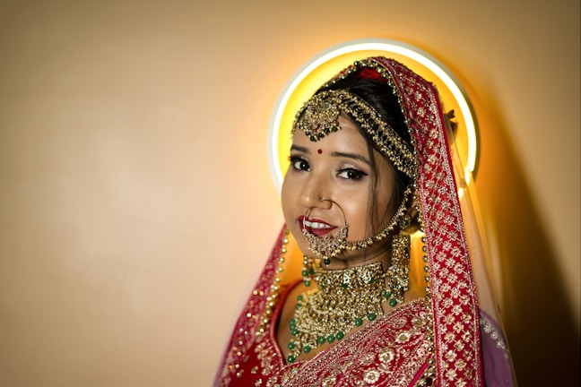 A bride adorned in traditional attire with intricate gold jewelry, including a nose ring, earrings, and a heavily embroidered red dupatta. Her makeup is elegantly done, featuring a bindi on her forehead. A halo-like circular light creates a soft glow behind her head.
