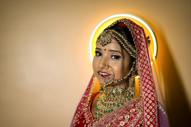 A bride adorned in traditional attire with intricate gold jewelry, including a nose ring, earrings, and a heavily embroidered red dupatta. Her makeup is elegantly done, featuring a bindi on her forehead. A halo-like circular light creates a soft glow behind her head.