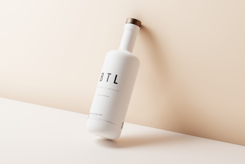A sleek, minimalist bottle is positioned diagonally against a soft beige background. The bottle is matte white with a simple label featuring large black letters 'BTL'. A wooden cap adds a touch of natural texture, contrasting with the bottle's smooth surface.