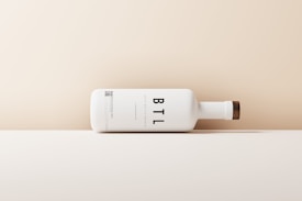 A minimalist, horizontally positioned white matte bottle with a wooden cap design. Large black letters 'BTL' are prominently displayed on the side. The background features a gradient of beige to cream, creating a clean, modern aesthetic.