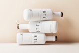 Three white bottles are artistically stacked on top of each other against a light beige background. The bottles have a minimalist design with the text 'BTL' prominently displayed in large, bold letters. Each bottle has a wooden cap, giving a natural and elegant touch to the overall appearance.