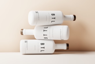 Three white bottles are artistically stacked on top of each other against a light beige background. The bottles have a minimalist design with the text 'BTL' prominently displayed in large, bold letters. Each bottle has a wooden cap, giving a natural and elegant touch to the overall appearance.