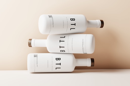 Three white bottles are artistically stacked on top of each other against a light beige background. The bottles have a minimalist design with the text 'BTL' prominently displayed in large, bold letters. Each bottle has a wooden cap, giving a natural and elegant touch to the overall appearance.
