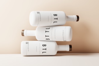 Three white bottles are artistically stacked on top of each other against a light beige background. The bottles have a minimalist design with the text 'BTL' prominently displayed in large, bold letters. Each bottle has a wooden cap, giving a natural and elegant touch to the overall appearance.