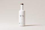 A tall white bottle with a minimalistic design is centered against a light gray background. The bottle features bold black letters spelling 'BTL' and additional smaller text below. It has a round wooden cap.