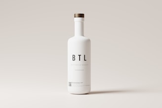 A tall white bottle with a minimalistic design is centered against a light gray background. The bottle features bold black letters spelling 'BTL' and additional smaller text below. It has a round wooden cap.
