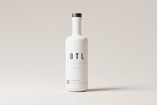 A tall white bottle with a minimalistic design is centered against a light gray background. The bottle features bold black letters spelling 'BTL' and additional smaller text below. It has a round wooden cap.