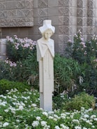 A stylized sculpture of a human figure stands amidst a lush garden filled with green foliage and white flowers. The backdrop features an intricately patterned, textured wall that adds a geometric element to the scene.