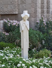 A stylized sculpture of a human figure stands amidst a lush garden filled with green foliage and white flowers. The backdrop features an intricately patterned, textured wall that adds a geometric element to the scene.