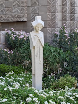 A stylized sculpture of a human figure stands amidst a lush garden filled with green foliage and white flowers. The backdrop features an intricately patterned, textured wall that adds a geometric element to the scene.