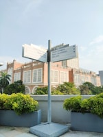 A directional signpost stands prominently in an outdoor setting, surrounded by lush greenery in planters. In the background, a large building with intricate architectural details and a red brick facade is visible under a partly cloudy sky.