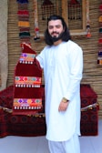 A man with long hair and a beard is wearing a white traditional outfit, standing in front of a textured background. He is holding a red velvet garment decorated with colorful embroidery and dangling beads. The background includes ornate, woven decorations and a cushioned bench covered in a patterned fabric.