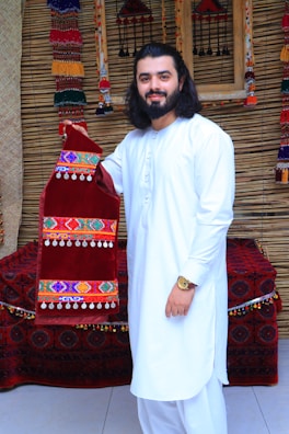 A man with long hair and a beard is wearing a white traditional outfit, standing in front of a textured background. He is holding a red velvet garment decorated with colorful embroidery and dangling beads. The background includes ornate, woven decorations and a cushioned bench covered in a patterned fabric.