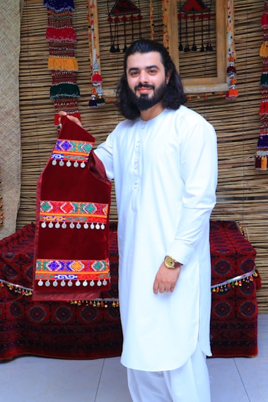A man with long hair and a beard is wearing a white traditional outfit, standing in front of a textured background. He is holding a red velvet garment decorated with colorful embroidery and dangling beads. The background includes ornate, woven decorations and a cushioned bench covered in a patterned fabric.