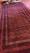 A richly patterned red carpet with intricate designs featuring geometric and floral motifs. The carpet displays a mix of deep red and dark colors with a series of repeated circular patterns. A small part of the edge is folded back, revealing the reverse side. Bare feet are visible at the top edge, stepping on the carpet.