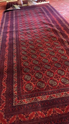 A richly patterned red carpet with intricate designs featuring geometric and floral motifs. The carpet displays a mix of deep red and dark colors with a series of repeated circular patterns. A small part of the edge is folded back, revealing the reverse side. Bare feet are visible at the top edge, stepping on the carpet.