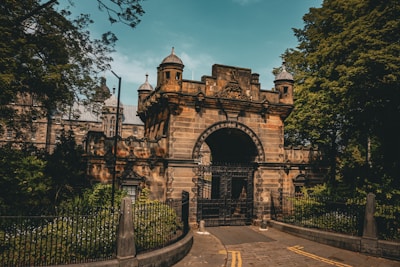 An ornate stone gate with intricate architectural details set within lush green surroundings. The gate features tall, cylindrical turrets with pointed domes, and is framed by wrought iron fencing. A large arch with decorative elements adds grandeur to the entrance. Behind the gate, there is a glimpse of a historic building with stone walls and tall windows. Overhanging branches and greenery enhance the peaceful, historic atmosphere.