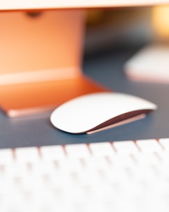 A close-up view of a modern, sleek computer mouse resting on a dark blue surface. The scene captures part of a desk setup with the edge of a keyboard in the foreground. The background is softly focused, featuring warm orange and gold tones, creating a cozy atmosphere.