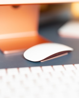 A close-up view of a modern, sleek computer mouse resting on a dark blue surface. The scene captures part of a desk setup with the edge of a keyboard in the foreground. The background is softly focused, featuring warm orange and gold tones, creating a cozy atmosphere.