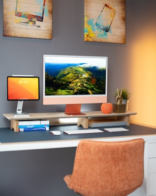 A modern home office setup features a large desktop monitor displaying a lush landscape scene with hills and greenery. A tablet displaying the time sits to the left, elevated by a stand. The desk is organized with a keyboard, mouse, and a stack of books. Two pieces of modern art depicting technology devices hang on the gray wall. An orange desk chair and a small plant add a touch of warmth to the space.