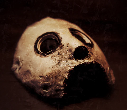 Dark, shadowy mask with deep creases and a sinister, snarling mouth revealing rough paper teeth.