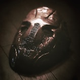 An eerie, metallic mask lies on a wooden surface, featuring a dark, distressed patina. The mask has a stitched design down the center, creating a haunting appearance with hollow eyes and indented facial features, evoking a sense of mystery and unease.