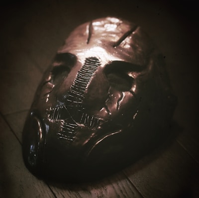 An eerie, metallic mask lies on a wooden surface, featuring a dark, distressed patina. The mask has a stitched design down the center, creating a haunting appearance with hollow eyes and indented facial features, evoking a sense of mystery and unease.