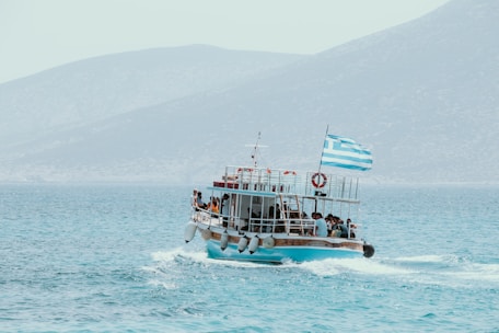 A boat filled with passengers travels across a calm sea with mountains in the background. A Greek flag waves at the stern of the boat, and several people are gathered on deck, enjoying the ride.